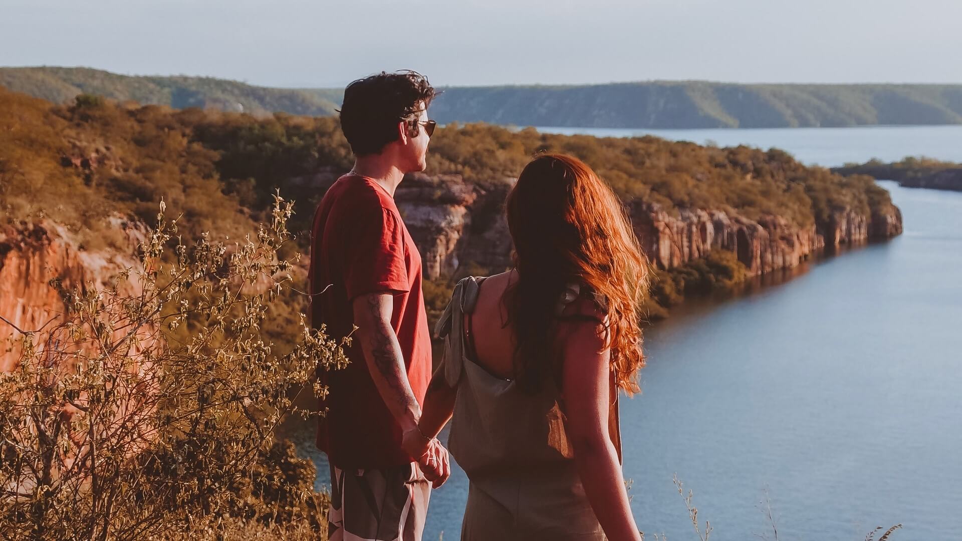 Homem e mulher de mãos dadas contemplam paisagem de rio cercado por cânions em dia ensolarado