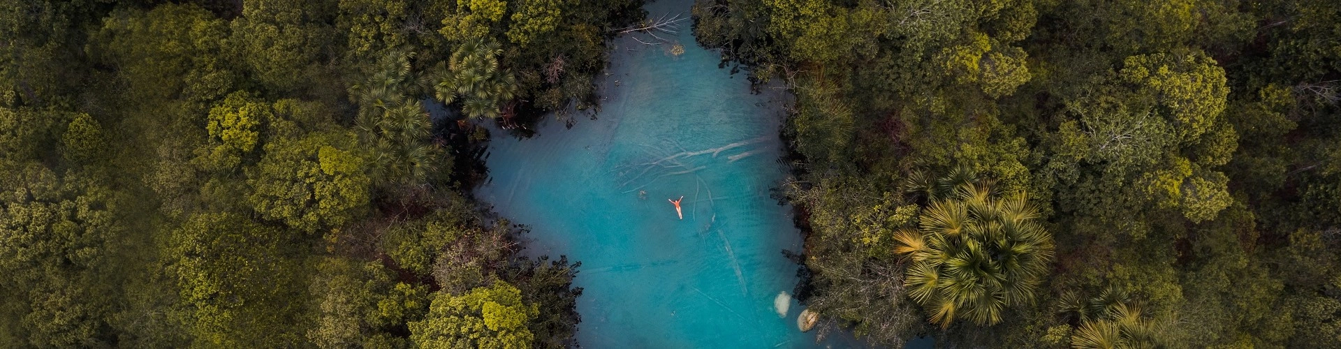 Vista panorâmica de uma lagoa de água azul cristalina onde uma pessoa flutua na superfície das águas com os braços abertos. Ao redor, várias árvores de folhas verdes cercam a vegetação.