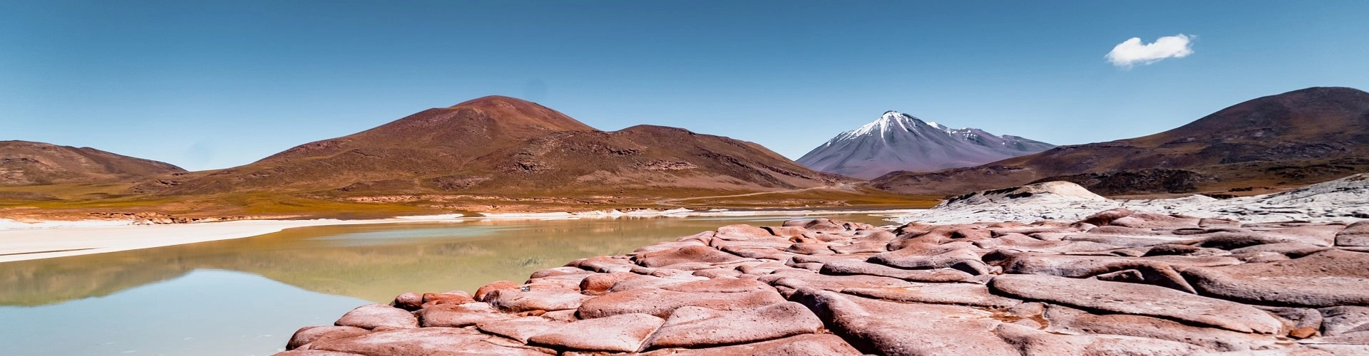 Cenário de muitas rochas, com montanhas e vulcão em segundo plano. No primeiro, ao lado do solo pedregoso e de cor  avermelhada, um riacho raso.