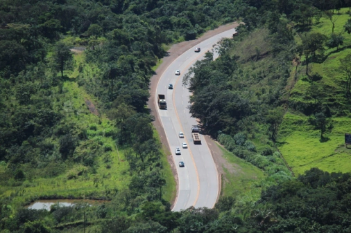 Vista aérea de trecho de estrada asfaltada, com curvas e cercada por vegetação. Alguns carros de passeio e caminhões estão na estrada