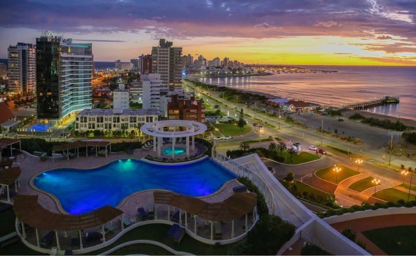 Vista aérea de cenário urbano em contraste com o pôr do sol refletindo na praia. No plano frontal, uma enorme piscina com as bordas curvadas e prédios ao fundo