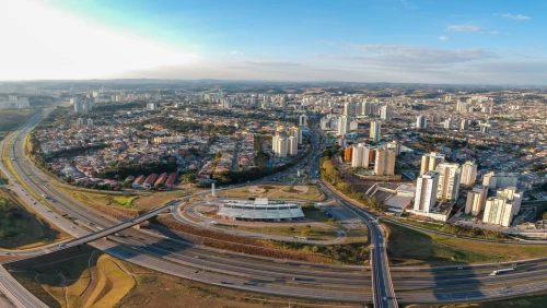 Vista aérea de uma paisagem urbana com estradas, ruas, edifícios e demais construções na cidade de Jundiaí, interior de SP. A foto ampla destaca o céu azulado e os bairros em expansão no entorno do município.
