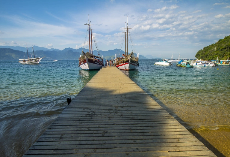Marina em praia de Angra dos Reis, com destaque para dois barcos ancorados, iates ao fundo, montanhas e céu azulado