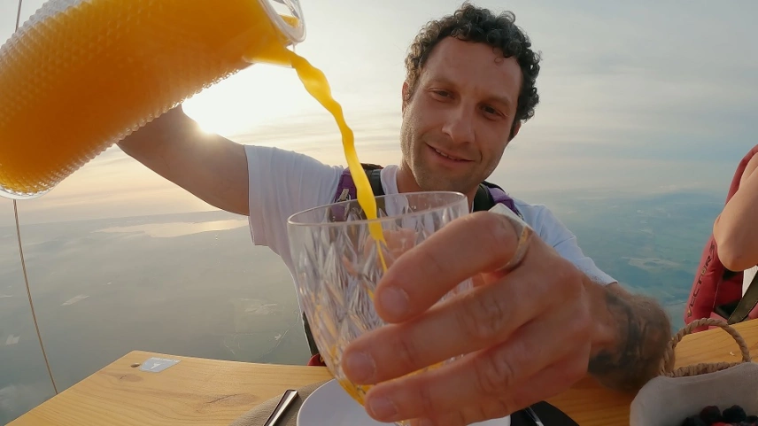 Um homem servindo um suco de cor laranja em uma mesa suspensa nos céus por um balão de ar quente.