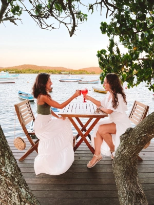 Duas mulheres sentadas em mesa de bar à beira da praia brindando com bebidas.