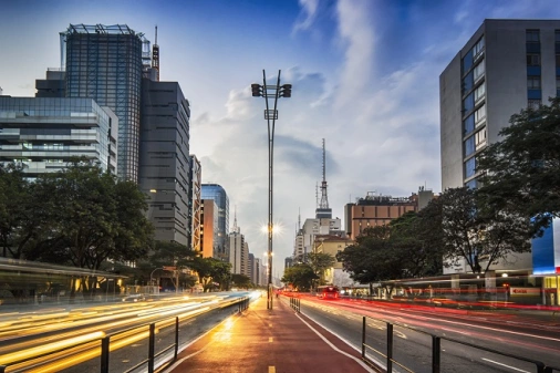 As ruas asfaltadas e vazias da Avenida Paulista, em São Paulo, com prédios e algumas árvores ao redor sob o céu que começa a escurecer com a chegada da noite