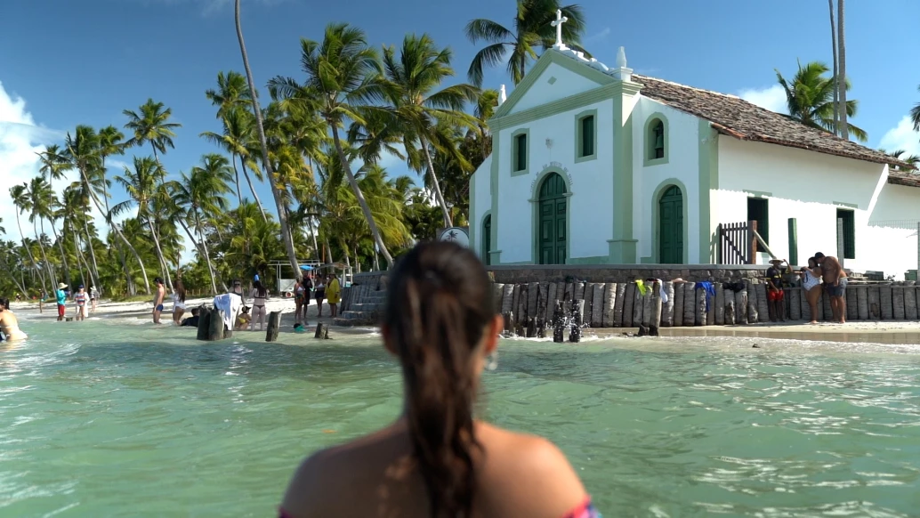 Mulher de costas para a câmera enquanto observa a fachada de uma igreja na Praia dos Carneiros. Há muitos coqueiros e alguns turistas ao fundo