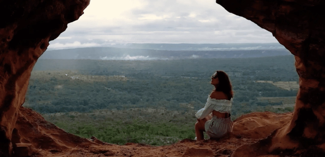 Abertura de formação rochosa vermelho-terrosa expõe um vasto horizonte verde. No canto, uma mulher de roupa branca aprecia a vista.