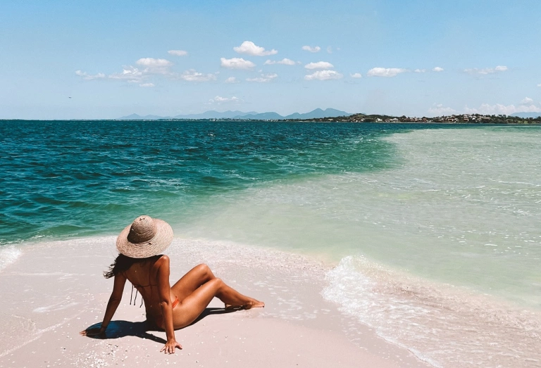 Mulher sentada em praia de areias claras avistando o verde mar e céu azul.