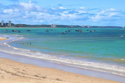 Paisagem da Praia de Pajuçara com águas verdes cristalinas cercada por prédios e montanhas ao fundo com céu azul