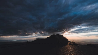 Feixes de luz solar entre nuvens marcando a silhueta de uma pedra e uma cruz no alto de uma colina