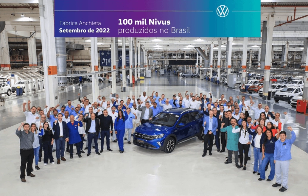 Dentro de uma fábrica da Volkswagen, ao redor de um Nivus azul, dezenas de funcionários celebram com as mãos para o alto.