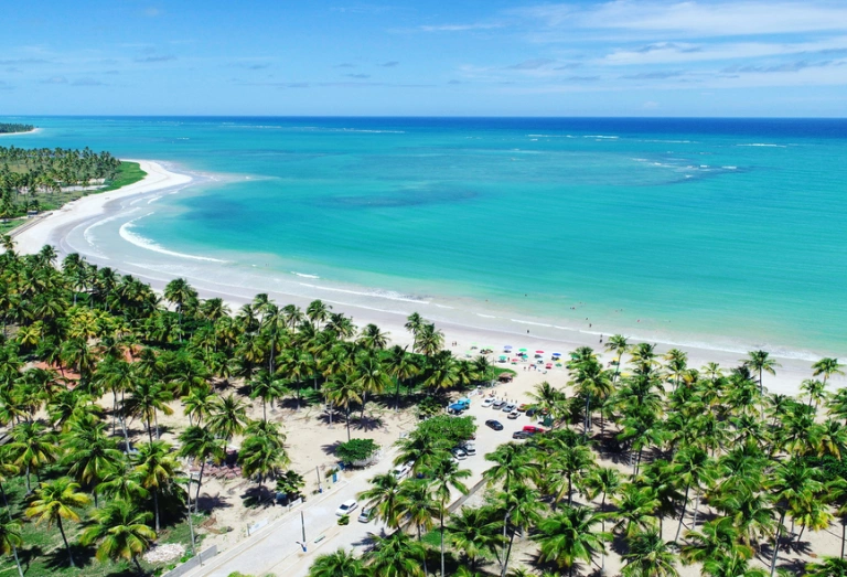Vista aérea de praia em São Miguel dos Milagres, Alagoas. O azul reluzente do lar contrasta com a areia branca e vegetação exuberante.