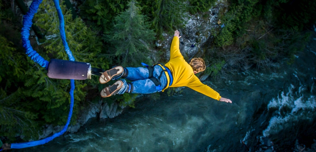 Homem branco e loiro, vestindo uma calça jeans e uma blusa amarela, salta de bungee jump sobre um rio com uma floresta às margens.