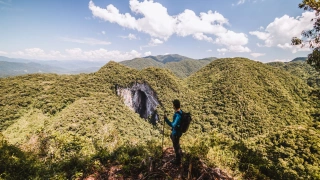 Homem em pé em um pico que dá vista para a enorme entrada de uma caverna. As montanhas ao redor são cobertas por vegetação densa