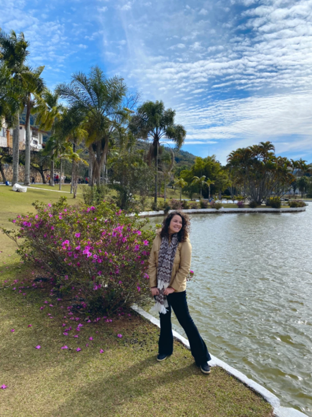 Mulher sorridente posando ao lado de um arbusto florido que na margem de um lago na Praça Adhemar de Barros, em Águas de Lindóia, SP. Ao fundo, grandes coqueiros e densa vegetação se contrastam com o céu azulado.