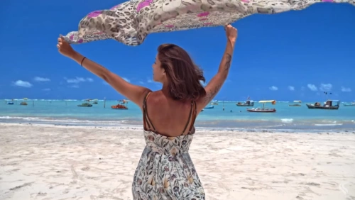 Mulher de costas levanta lenço em frente a praia de águas azuis em dia ensolarado