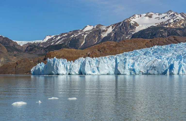 Enorme geleira de coloração azul na beira de um lago. Ao fundo, montanhas escuras com neve no topo