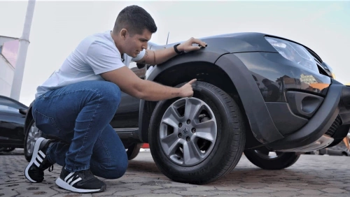 Agachado, o especialista em autos e influenciador Xenão aponta para o pneu de um carro.