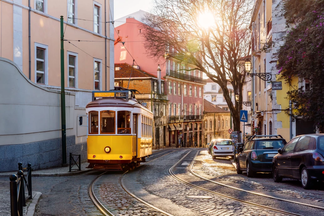 A imagem mostra um cenário pitoresco e ensolarado de uma rua em Portugal, onde se destaca um tradicional bonde amarelo. O bonde está em movimento ao longo dos trilhos que cortam a rua de paralelepípedos. Ao lado, alinhados na rua, há carros estacionados, o que pode remeter à conveniência do aluguel de carro em Portugal para explorar a cidade com flexibilidade. O sol baixo lança uma luz dourada sobre a cena, criando uma atmosfera acolhedora. Edifícios históricos com fachadas coloridas e detalhes arquitetônicos europeus caracterizam o ambiente urbano. Uma placa à direita indica o “Largo do Limoeiro”, proporcionando um senso de localização na imagem.