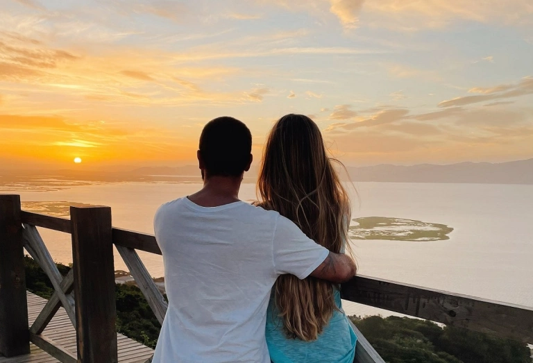 Casal de costas, abraçados, assistindo ao pôr do sol em um mirante com vista para o mar.