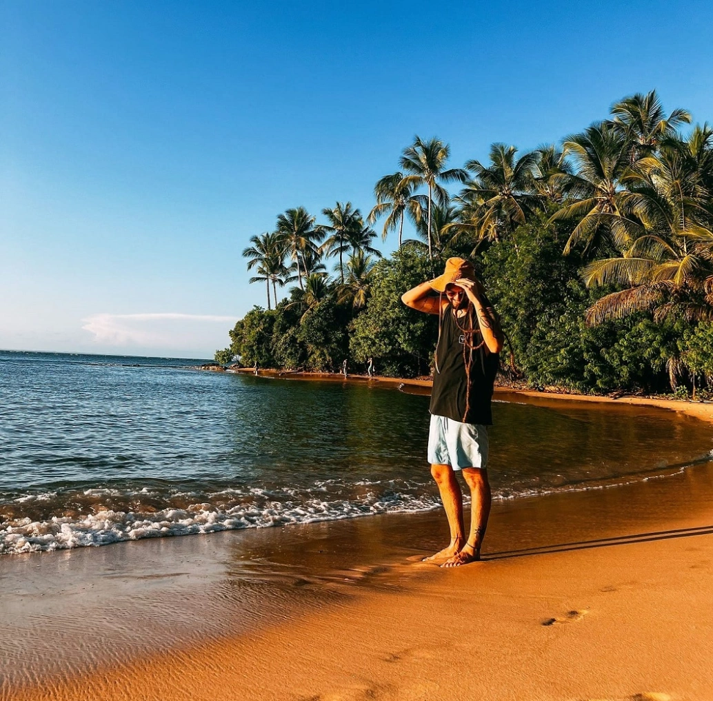 Homem em pé na beira da praia cercada por coqueiros em fim de tarde