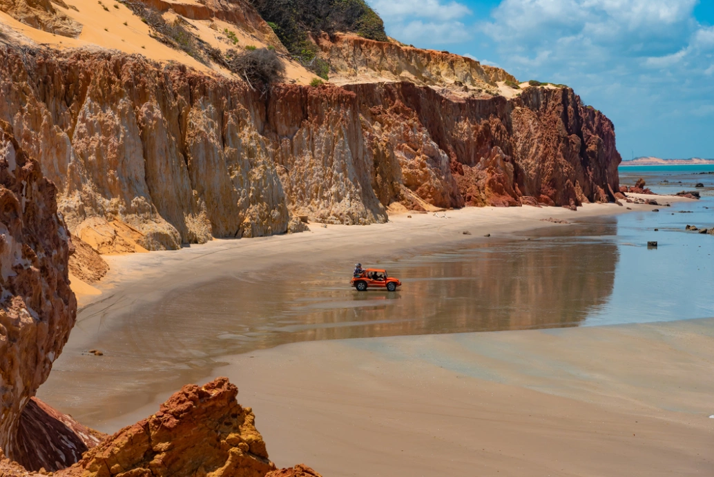 Vista lateral de falésias de cor alaranjada, algumas mais claras e outras mais escuras. Na areia da praia, um buggy com alguns turistas