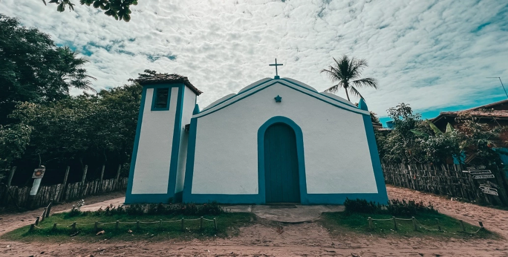 Fachada de pequena igreja nas cores branca e azul cercada por vegetação em dia claro