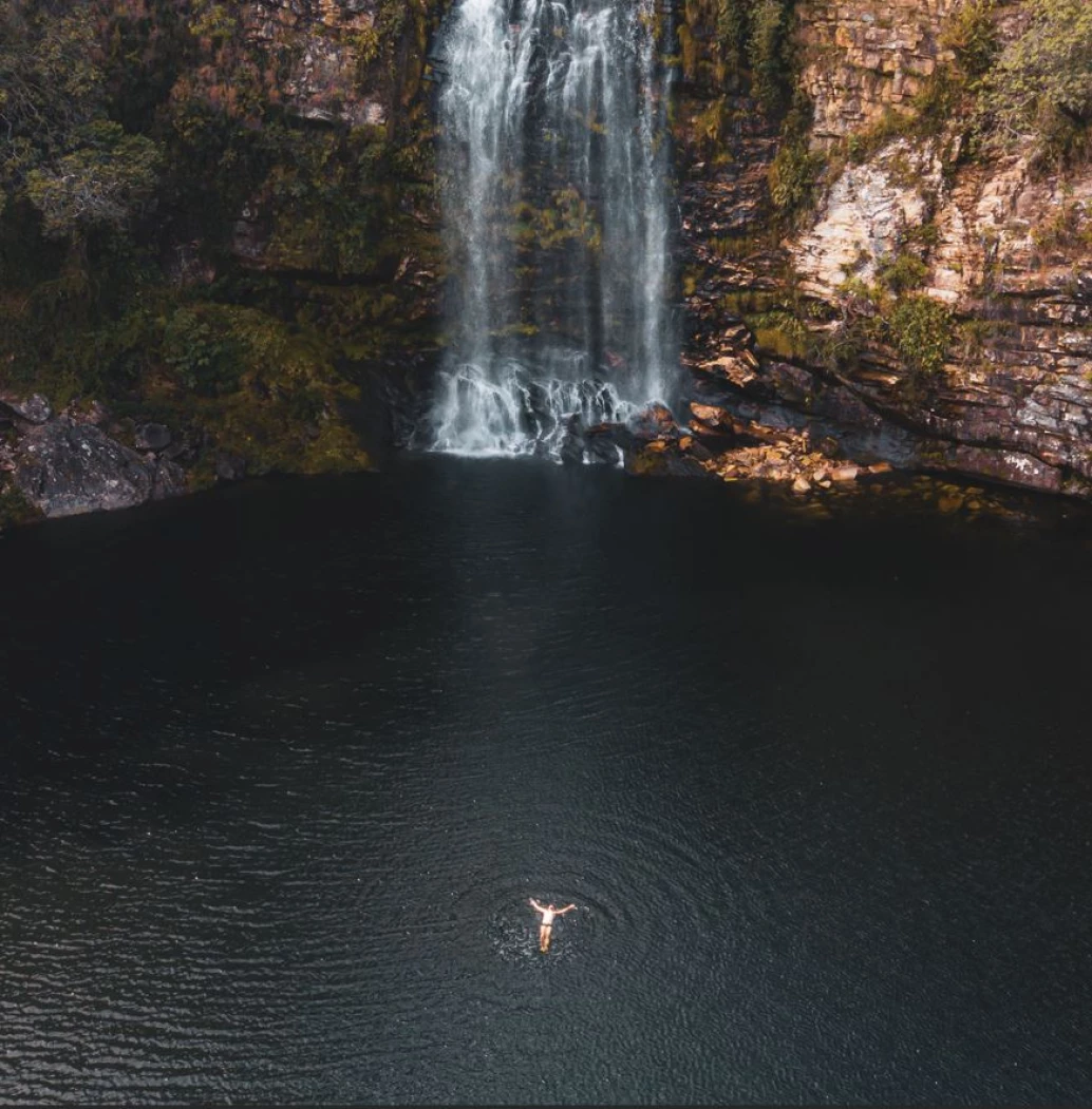 Vista aérea e distante de um homem flutuando com os braços abertos sobre o poço de uma cachoeira na Serra do Cipó. A queda d’água aparece ao fundo, em meio a um paredão rochoso parcialmente coberto por vegetação