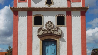 Fachada de igreja com parede branca, colunas vermelhas e porta azul em Lavras Novas. Há uma porteira azul no plano frontal, uma cruz no topo da igreja e céu azul com nuvens ao fundo