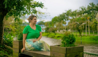 Mulher sorridente aprecia a vista sentada em banco de madeira em espaço aberto e arborizado durante o dia. O vibrante verde natural se destaca na imagem combinando com a roupa da mulher.