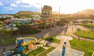 Vista panorâmica de parque de diversões em formato de castelo multicolorido com céu azulado ao fundo. Ao redor praças com grama verde e um helicóptero estacionado.