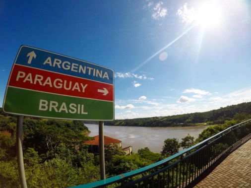 Foco em uma placa localizada na tríplice fronteira, indicando as direções para Argentina, Paraguai e Brasil. O cenário é um dia ensolarado, um rio e vegetação ao fundo