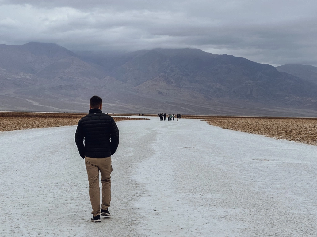 Homem de costas andando sobre um campo de sal. Ao fundo, um grupo de pessoas no mesmo caminho, enormes montanhas rochosas e céu coberto por nuvens e névoa.