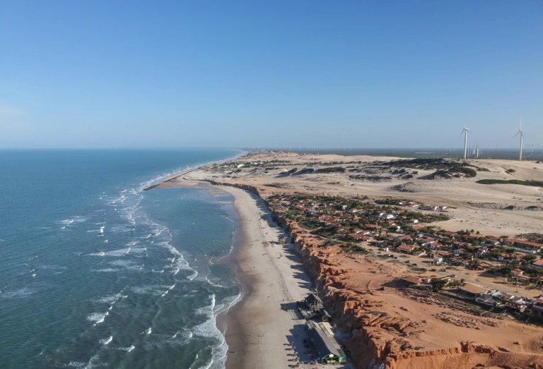 Vista aérea de praia com extensa faixa de areia, falésias avermelhadas e dunas de areia ao fundo