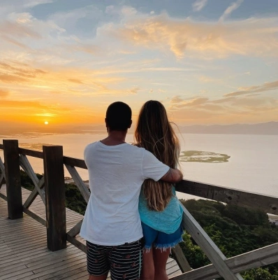 Homem e mulher assistem pôr do sol na praia em mirante