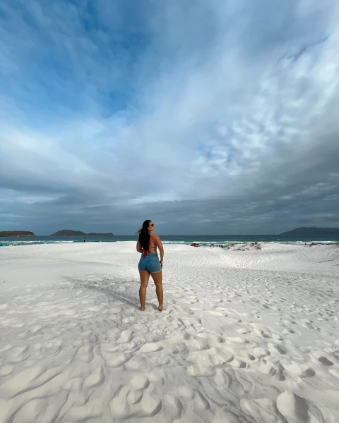 Mulher posando de costas para a câmera e de frente para o mar em uma praia de Cabo Frio. A areia é branca e fofa, formando pequenas dunas. O céu ao fundo está com algumas nuvens.