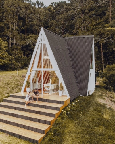 Casal sentado à frente de casa em formato triangular e com janelas de vidro em região arborizada
