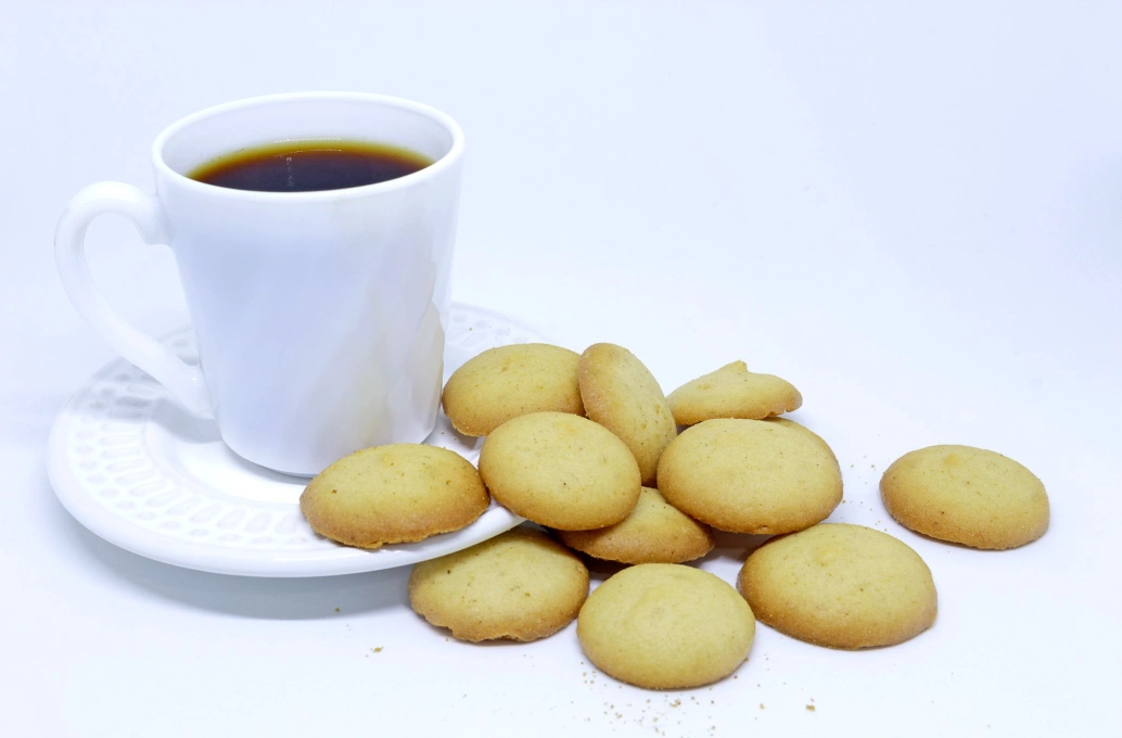 Bolinhos tareco, muito semelhantes com biscoitos,  espalhados por uma mesa branca e ao lado do pires e xícara brancos. A xícara está servida de café
