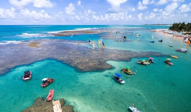 Vista aérea do mar verde-esmeralda e recifes de corais, com muitas jangadas sobre a água. Ao fundo, coqueirais, banhistas, guarda-sóis e céu azul com nuvens