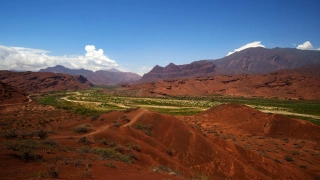 Visão de natureza parcialmente desértica, com montes e montanhas alaranjadas e um curso de vegetação verde ao centro. O céu azul limpo ao fundo contrasta com a paisagem