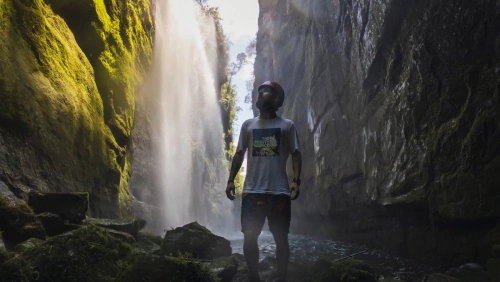 Fayson Merege em pé olhando para o alto na Cachoeira Véu da Noiva. Quedas d'água correm pelas paredes das rochas.