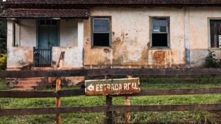 Na grade de madeira de uma velha fazenda, placa antiga e enferrujada escrito “Estrada Real”, apontando para a esquerda