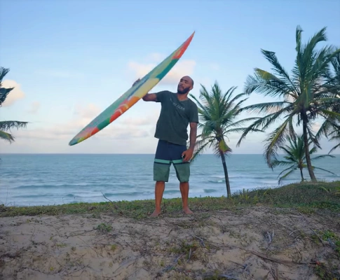 Homem segura prancha de surfe em frente à praia em dia claro