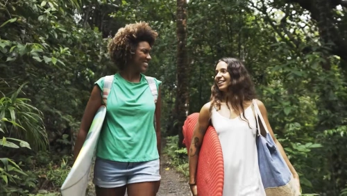Duas mulheres sorrindo uma para a outra carregam pranchas de surfe debaixo do braço descendo uma trilha cercada por árvores