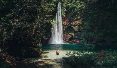 Clareira na mata em Barra das Garças, MT, revela uma linda cachoeira com piscina natural verde-esmeralda cercada de densa vegetação recebendo uma bracha de luz natural em dia claro. Ao centro da imagem, um jovem casal se abraça