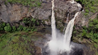 Vista aérea de uma imensa queda d’água que emerge de pedras e deságua em um poço. Vegetação ao redor da cachoeira