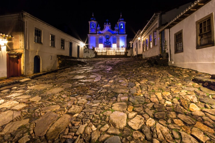 Vista noturna de igreja colonial em estilo barroco em cidade histórica de Minas Gerais. No plano frontal, destaque para a rua de pedra