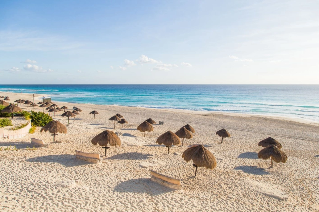 Bela praia na costa do Caribe. O mar azul ao fundo contrasta com a areia branca e os guarda sol de coqueiro.