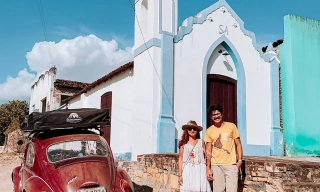Um homem e uma mulher em pé em frente a uma igreja com fachada branca em dia ensolarado com um Fusca vermelho estacionado ao lado